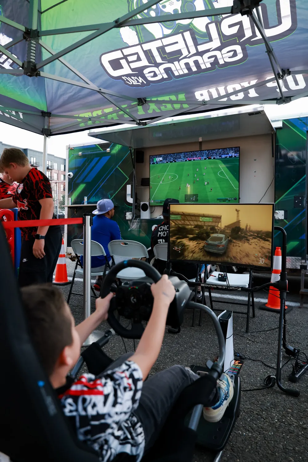 Kid on the racing simulator while guests play FIFA on the Uplifted Gaming trailer screen